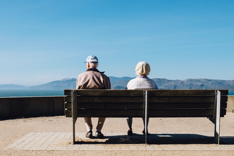 An elderly couple sitting on a bench together and looking out over mountains and the water