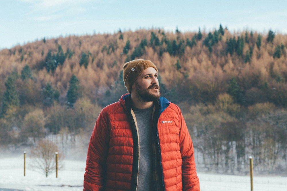 Man with a moustache and beard wearing a toque and a puffy jacket with snow and trees in the background
